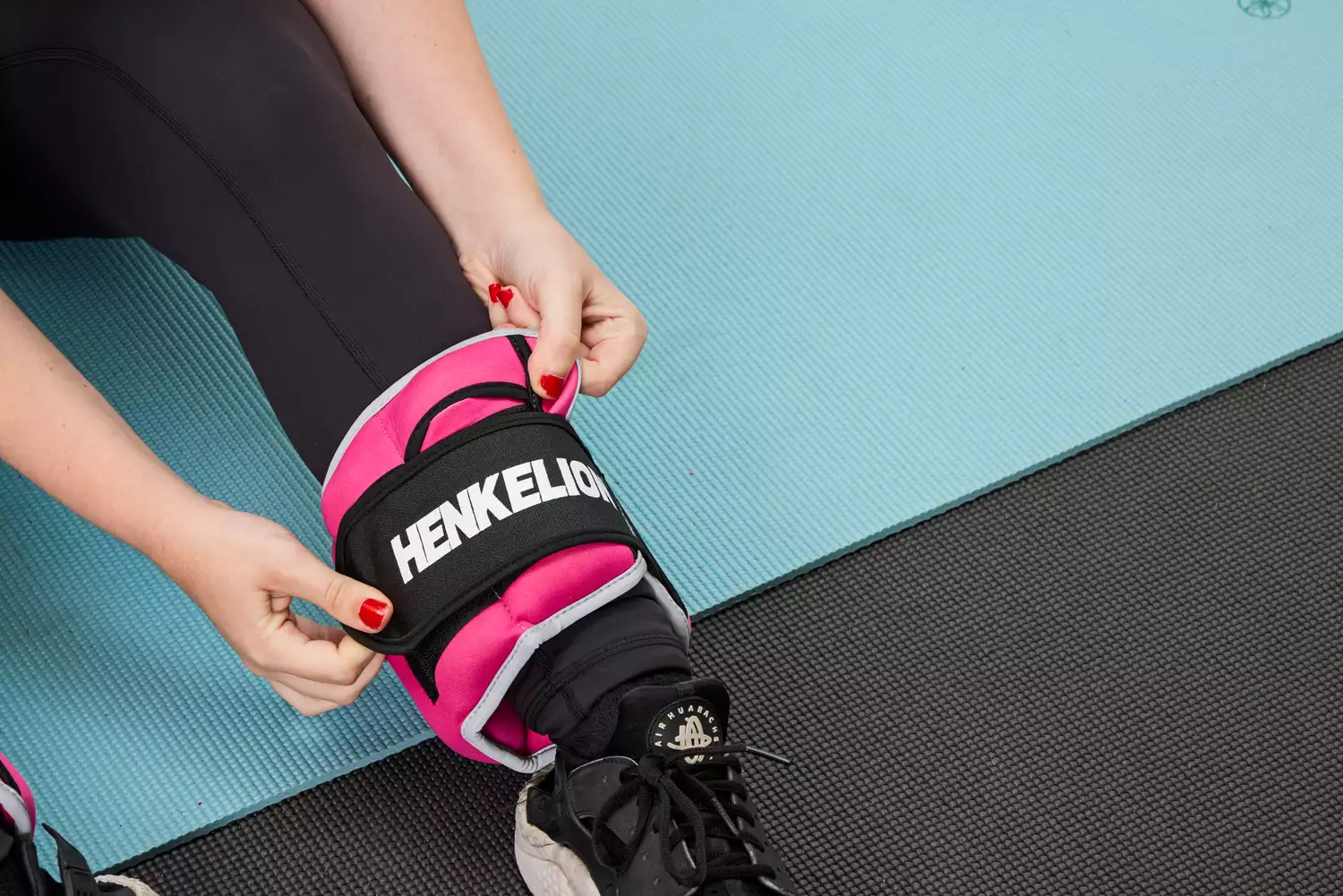 A person sitting on a yoga mat and securing a pink Henkelion ankle weight around their lower leg.