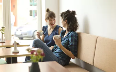 Two women laughing and enjoying coffee together in a brightly lit cafe booth.