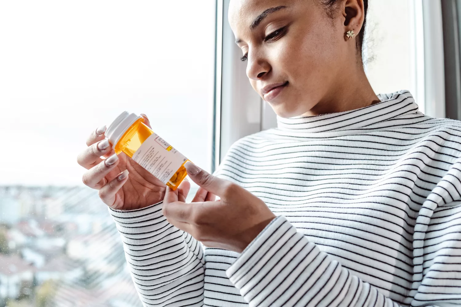 A woman thoughtfully looking at her medication bottle while standing in natural light.