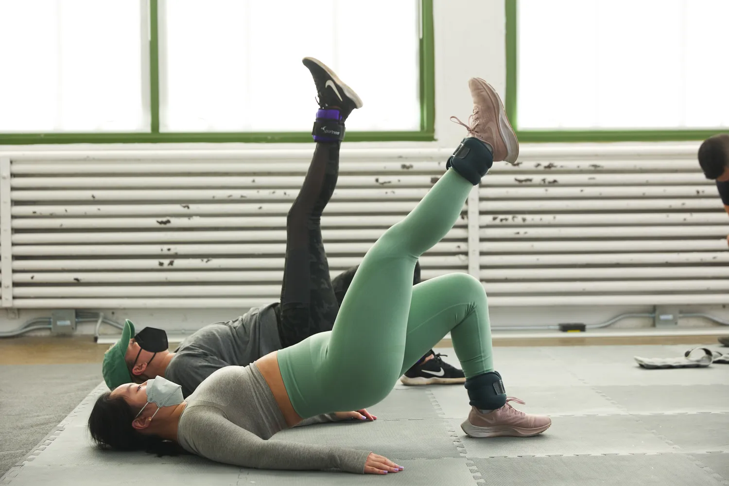 Two people performing leg-lift exercises with weighted ankle bands in a bright fitness studio.