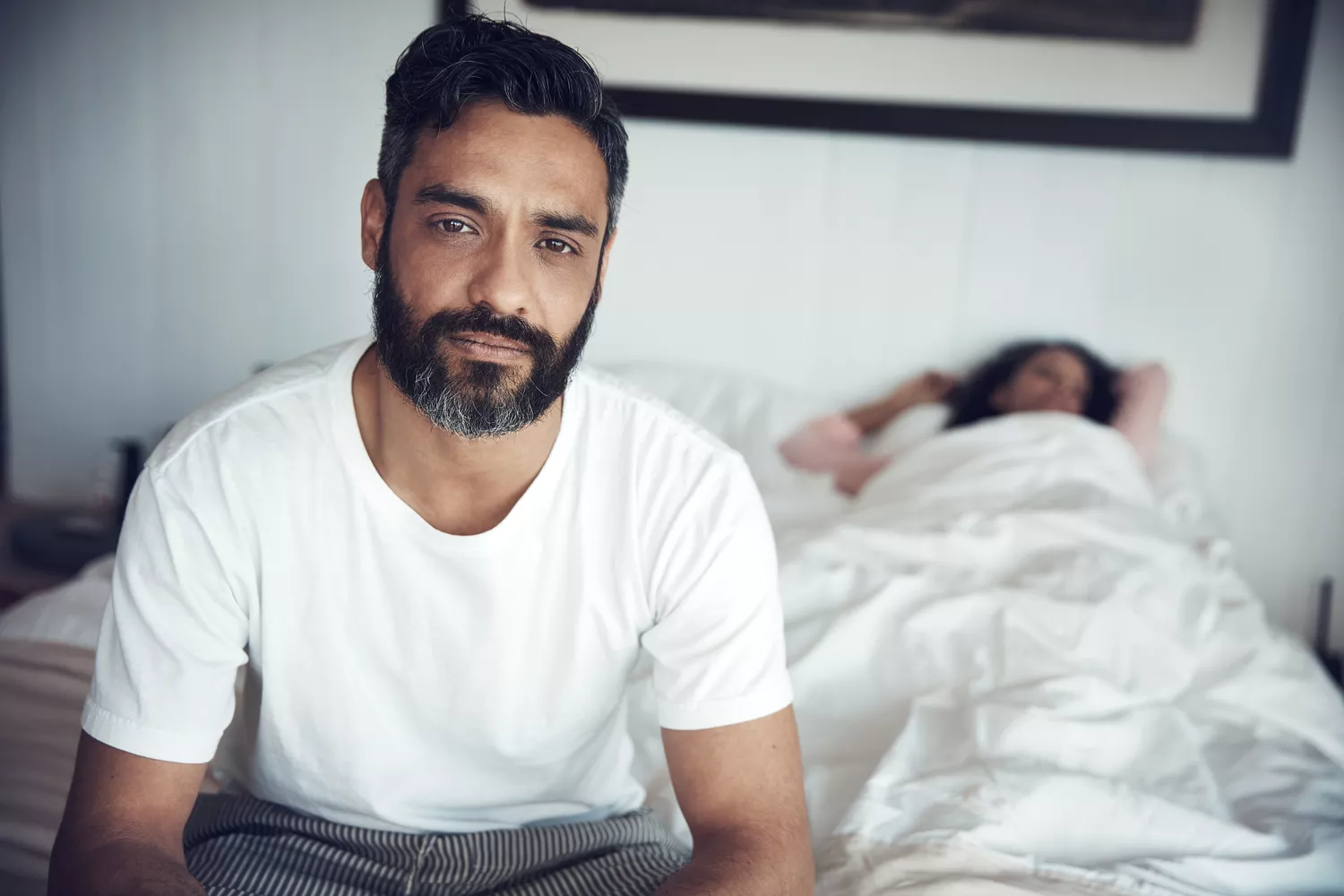 A couple sitting in bed looking frustrated or distant, depicting performance anxiety induced erectile dysfunction.