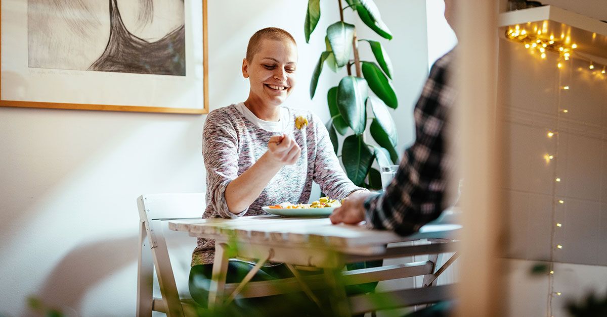 A woman with a shaved head smiling and enjoying a healthy meal at a table.
