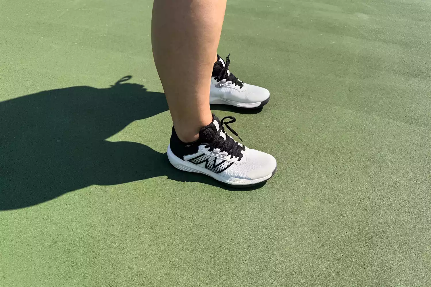 A person wearing New Balance tennis shoes on a green hardcourt, showing the wider base of the shoe.