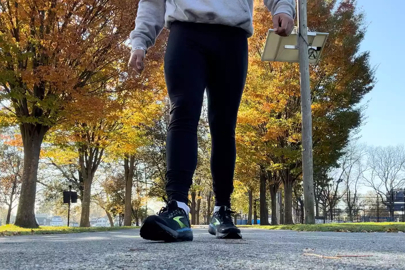 A runner in black leggings and a gray sweatshirt walking through a park with autumn leaves.