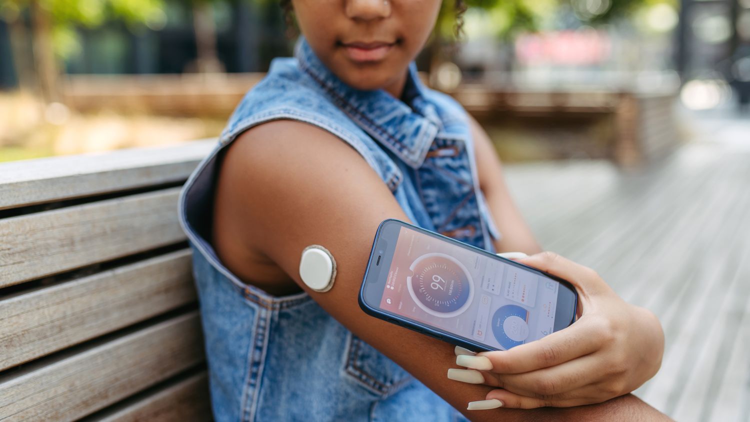 A woman checking her blood sugar levels on a smartphone using data from a continuous glucose monitor (CGM) while sitting on a park bench.