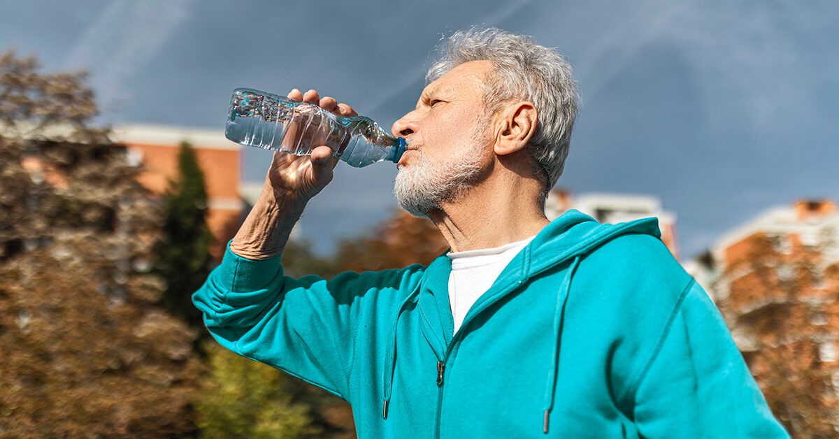 An older man in a teal hoodie drinking water outdoors during a workout session.