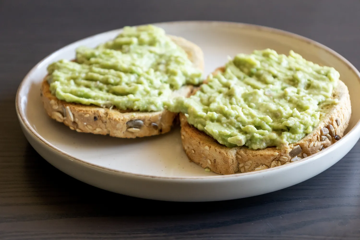 Two slices of seeded toast topped with mashed avocado on a white plate.