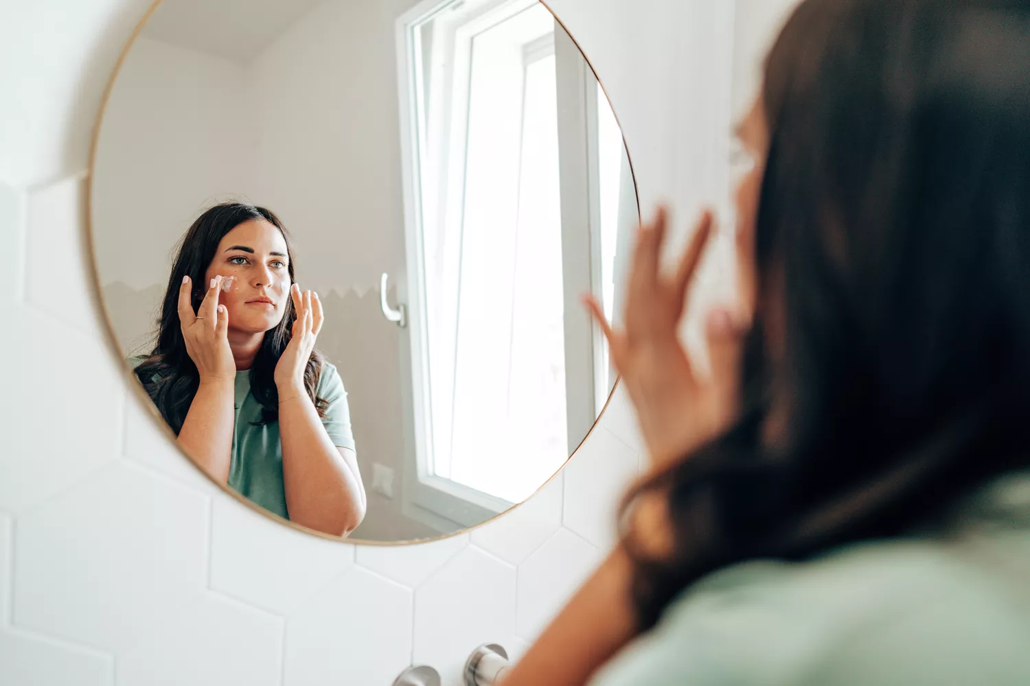 A woman looking at her reflection in a bathroom mirror while preparing for her day.