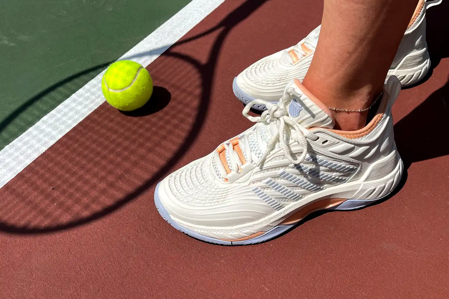 Close-up of tennis shoes on a red clay court next to a tennis ball.