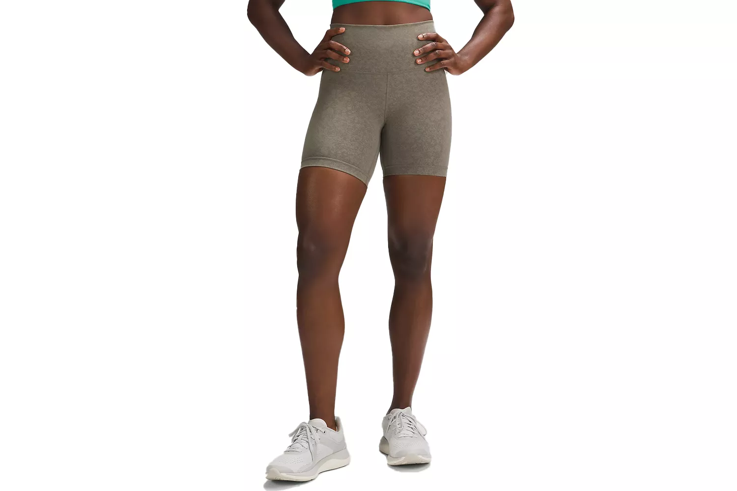 A model in olive green athletic shorts and a teal top posing in white sneakers.