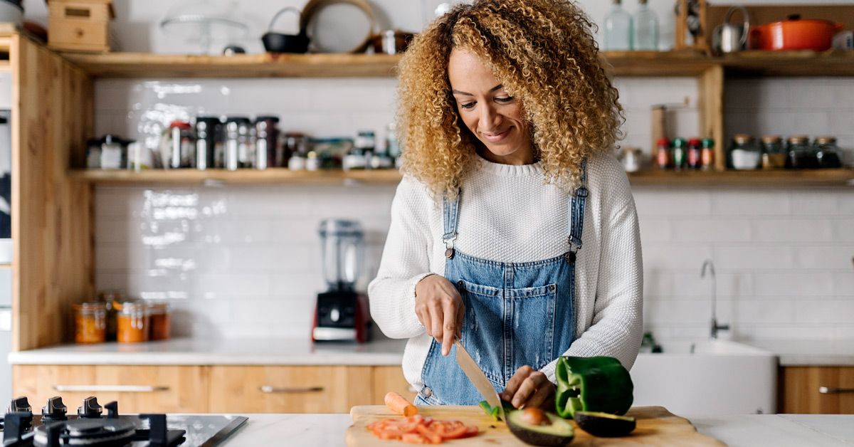 A woman with curly hair smiles while chopping fresh vegetables like avocado and bell pepper in a bright kitchen.