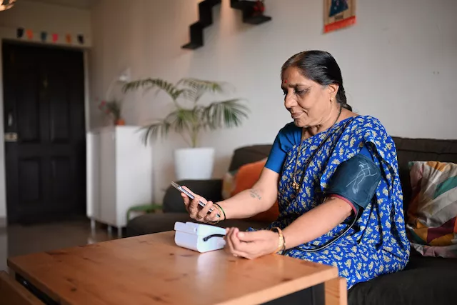 An older woman using a digital home blood pressure monitor while holding a smartphone.