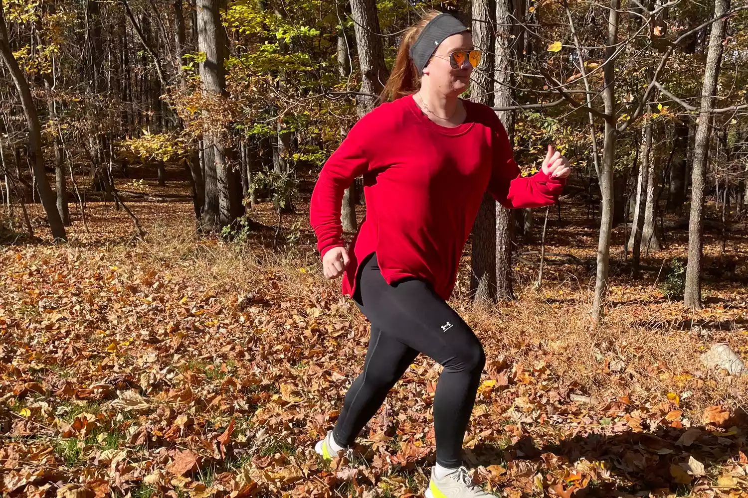 A woman running through a leaf-covered forest trail in black compression leggings and a red top.