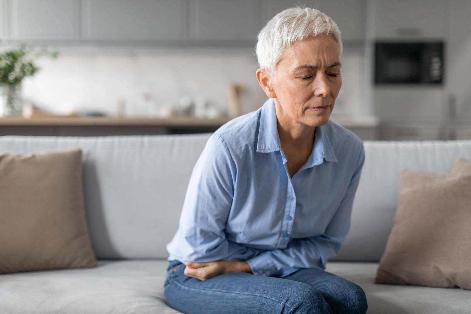 An elderly woman sitting on a sofa looking distressed and holding her stomach due to abdominal pain.