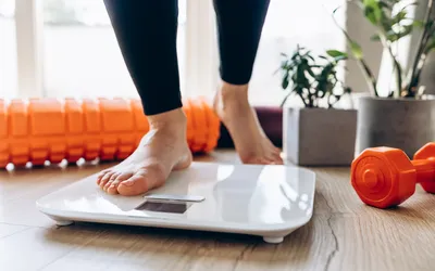 A person standing on a digital scale in a home gym environment with fitness equipment nearby.