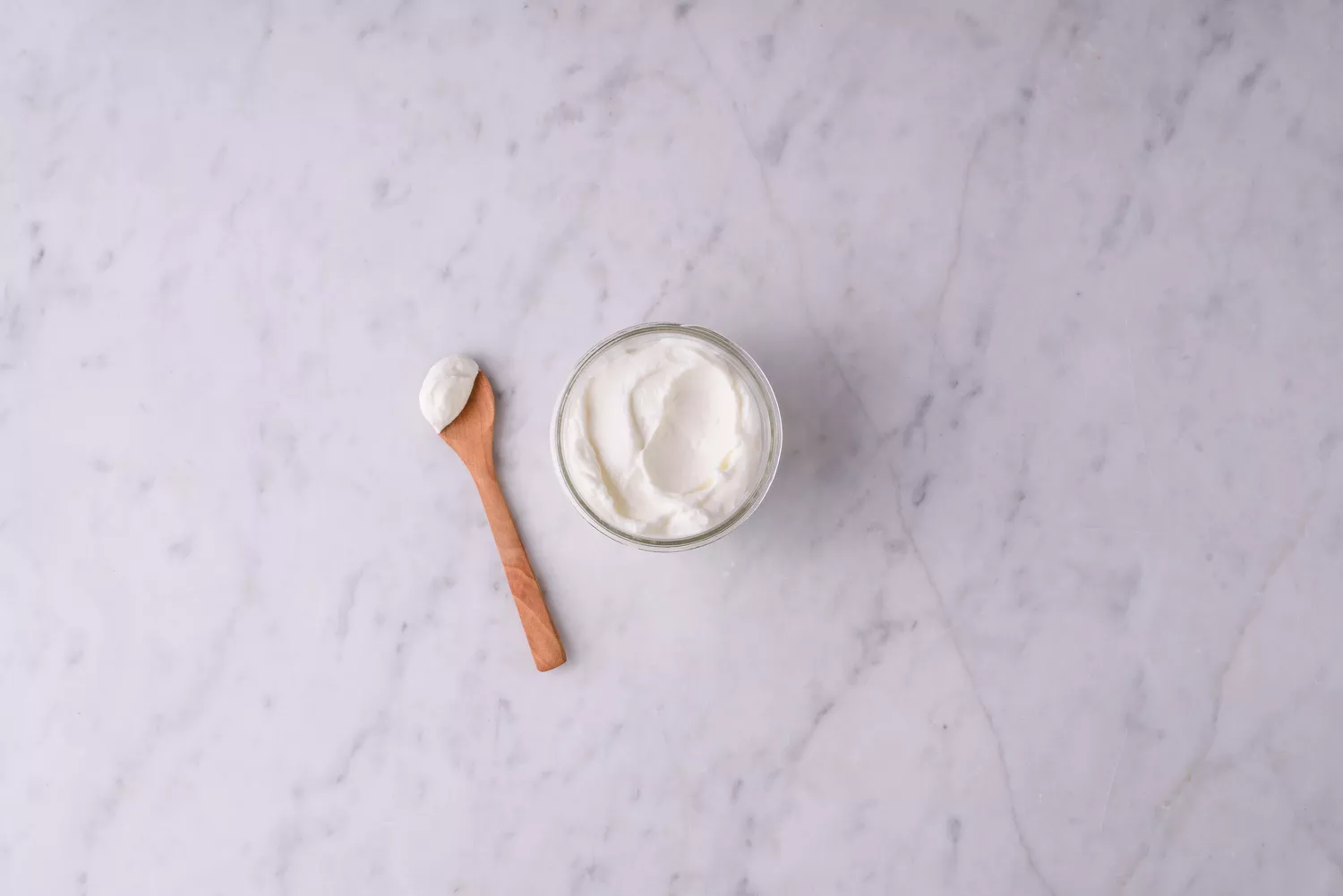 A glass jar of creamy white yogurt next to a wooden spoon on a marble surface.
