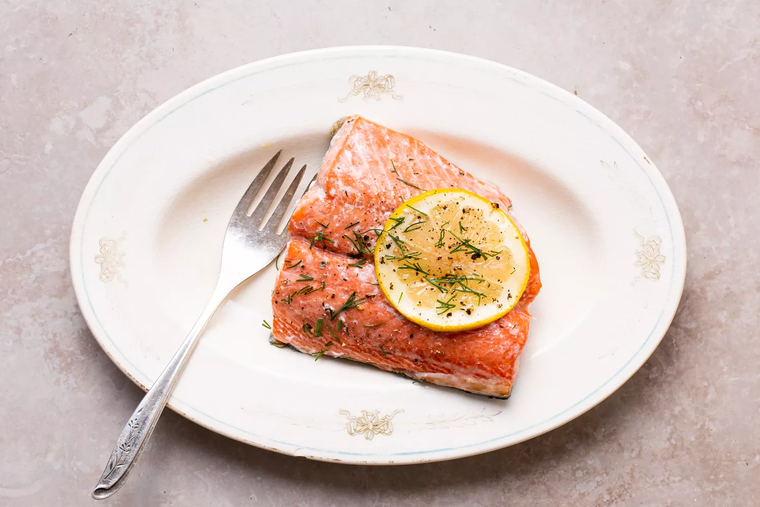 A cooked salmon fillet garnished with lemon and dill on a white plate.