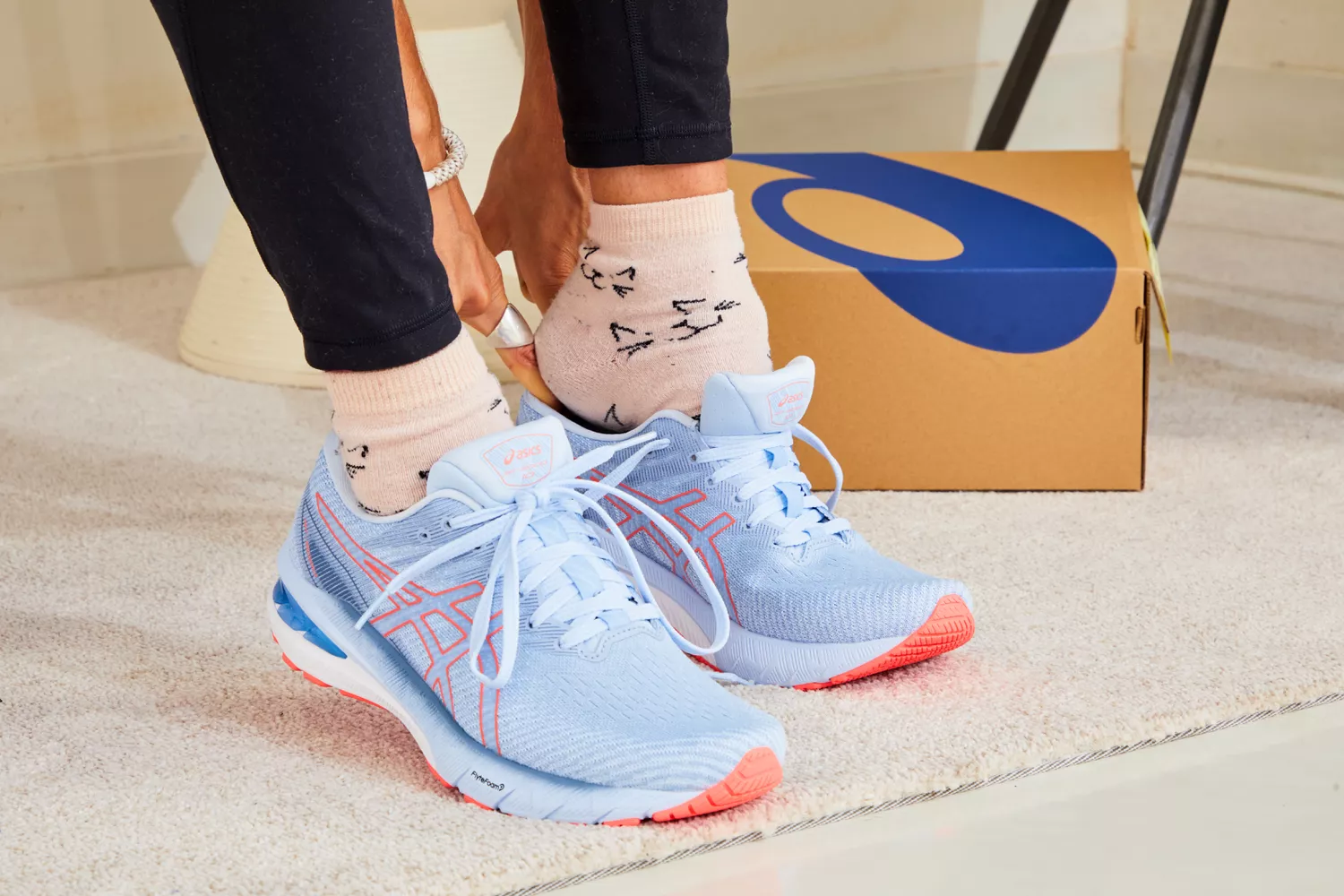 A person trying on a pair of light blue ASICS sneakers on a carpeted floor next to a shoebox.