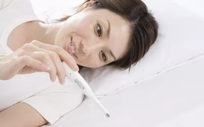 A woman lying in bed holding a digital thermometer, looking relaxed and focused on her health.