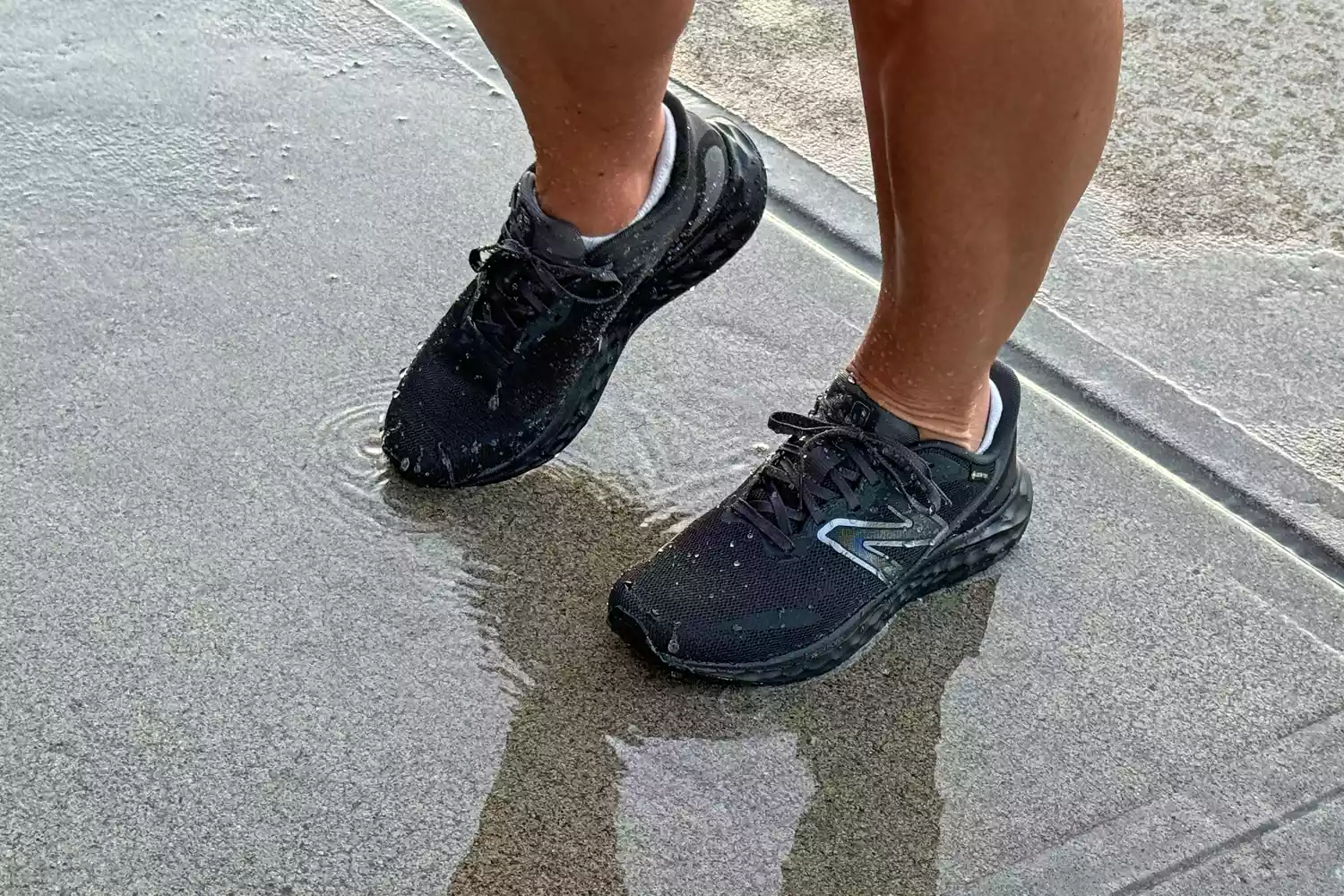 A person wearing black New Balance running shoes walking through a puddle on a sidewalk.