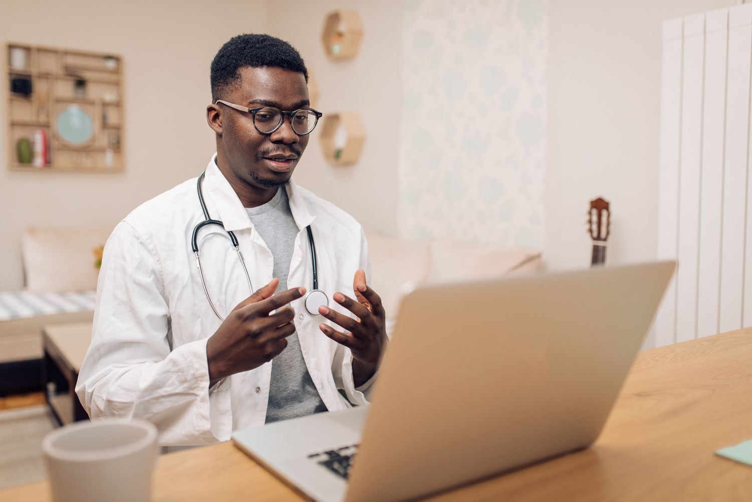A Black male doctor wearing a stethoscope talks to a patient through a laptop screen during a virtual health appointment.