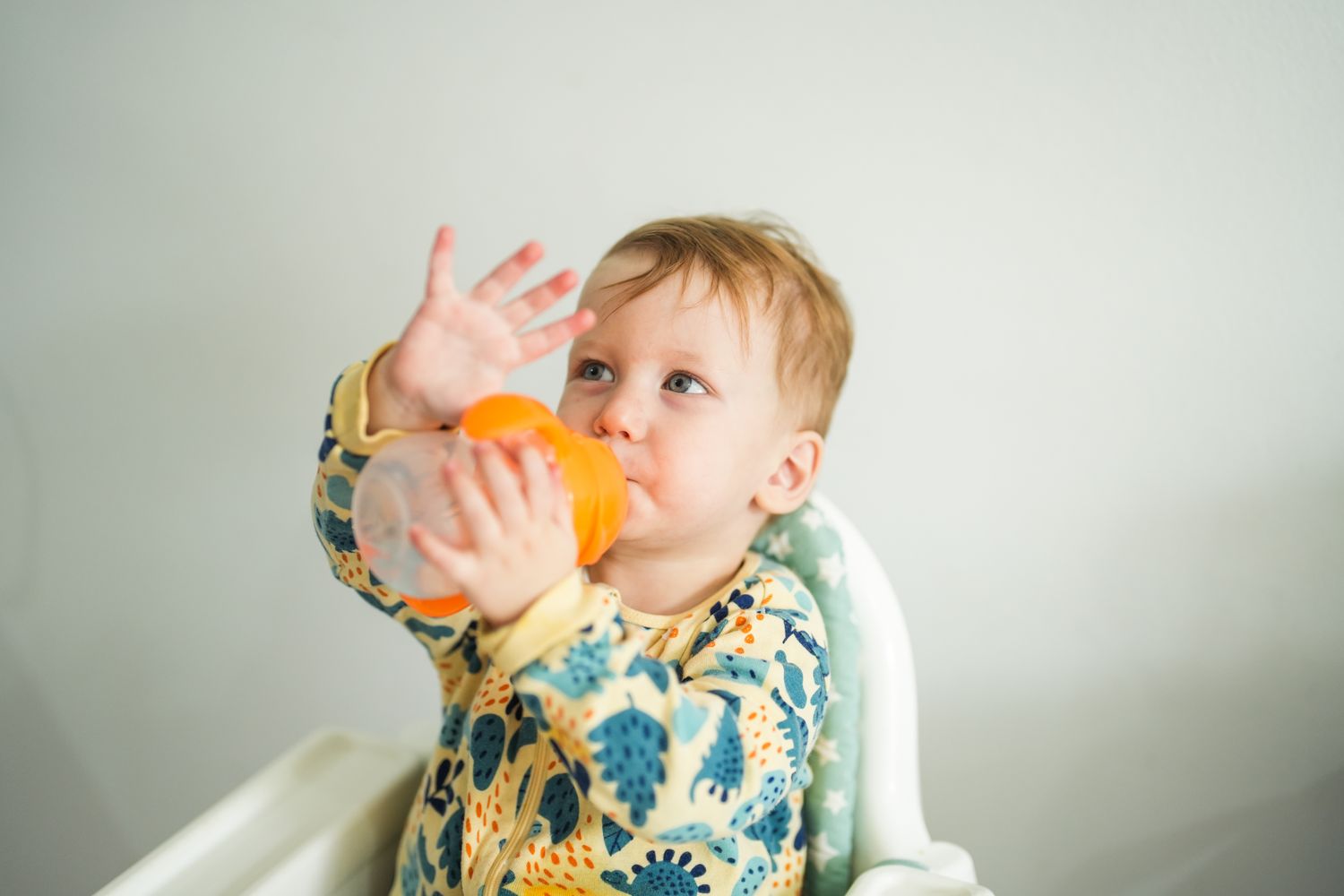A toddler in a dinosaur-print onesie drinking from a sippy cup.