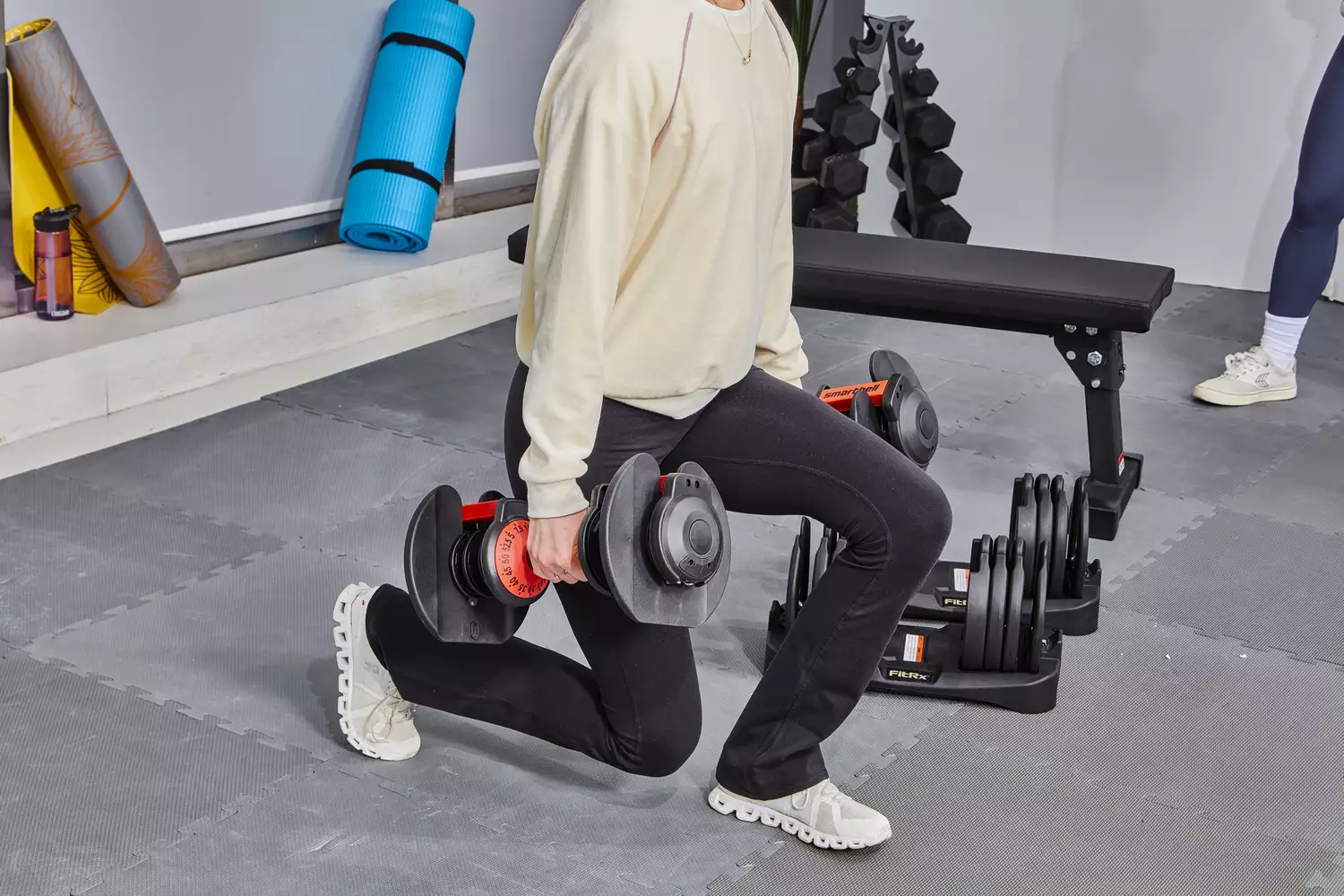A woman performing lunges with adjustable dumbbells in a home gym setting.