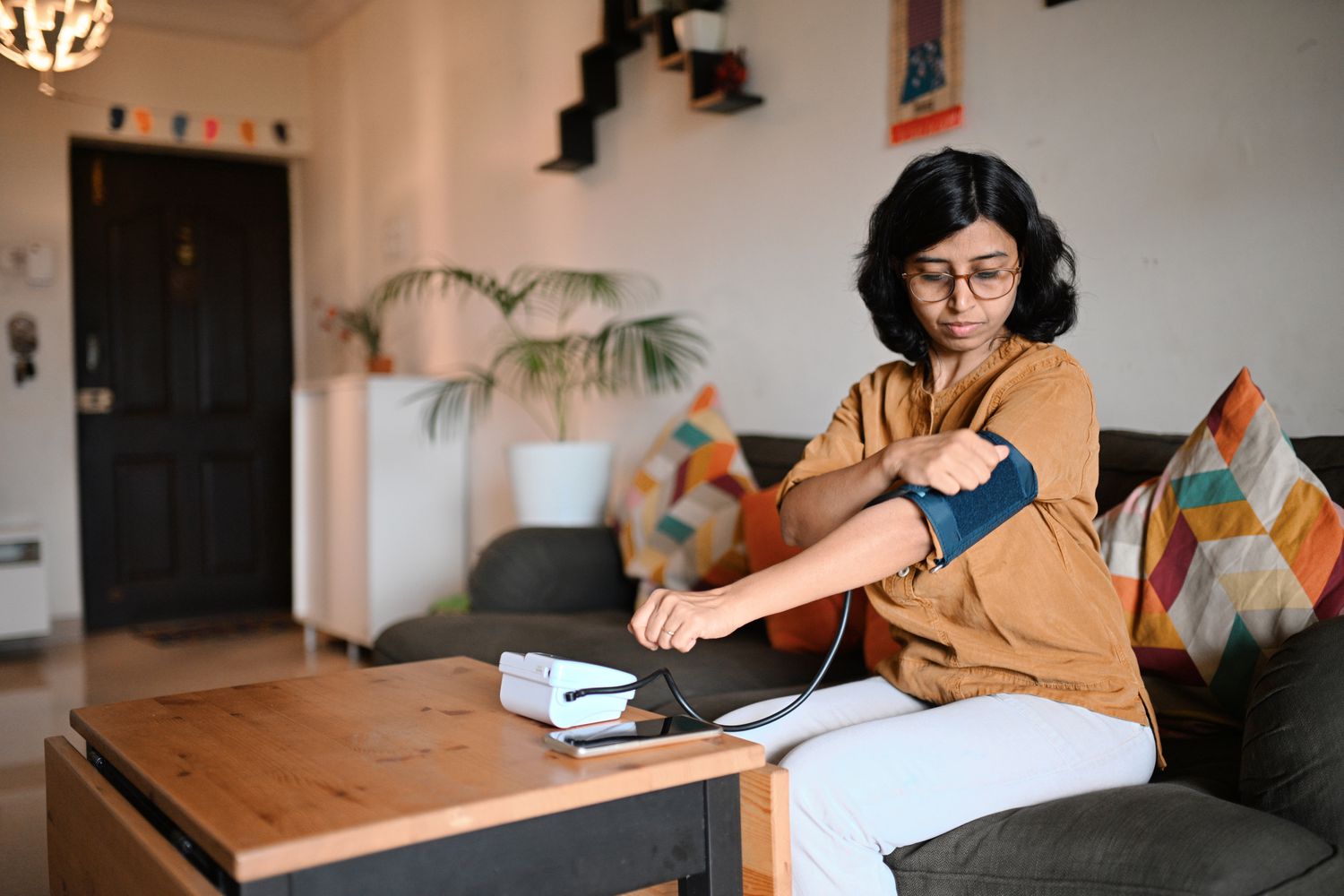 A woman sitting on a couch checking her blood pressure with a digital monitor while her phone sits on the table.
