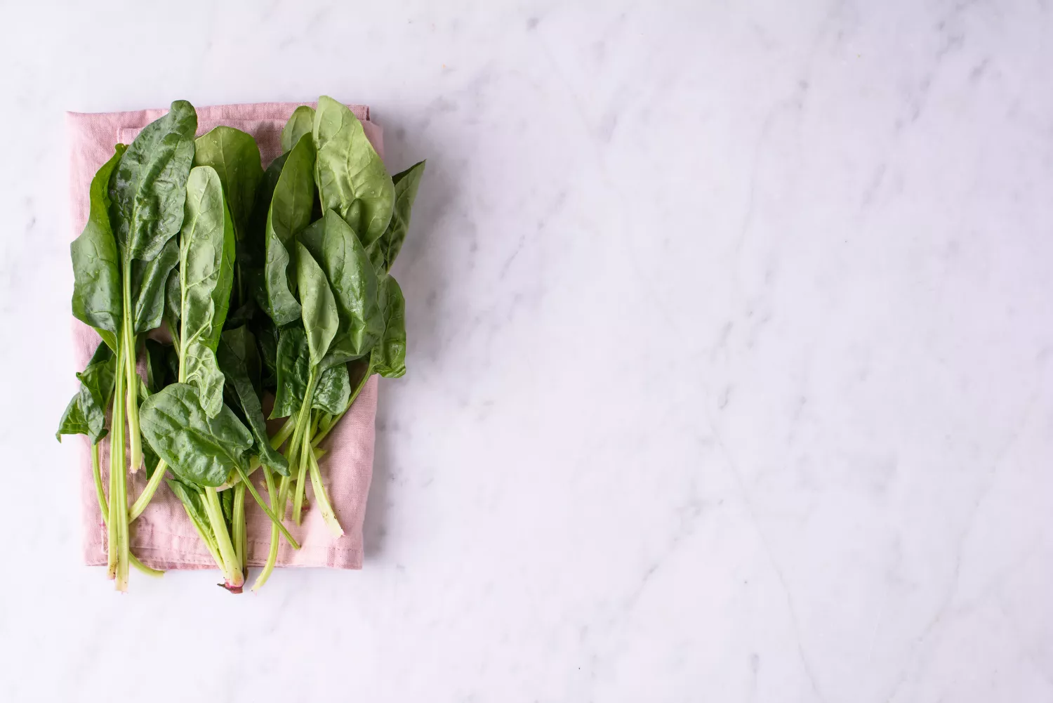 Fresh green spinach leaves arranged on a white marble surface with a pink cloth.