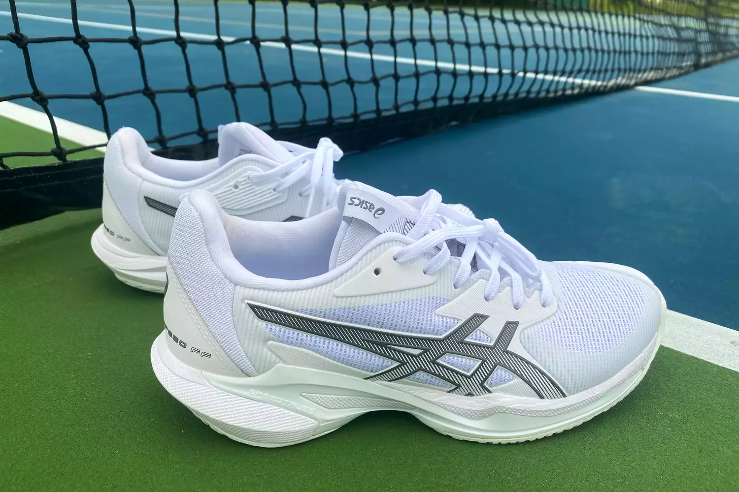White Asics Solution Speed FF 3 tennis shoes resting on a green court near the net.